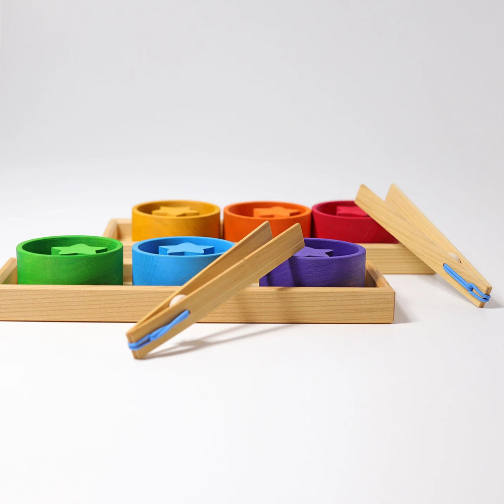 Grimm’s Sorting Game Rainbow Bowls with Tweezers featuring six colorful wooden bowls in a sorting tray, each containing a matching wooden star, with two wooden tweezers resting nearby for fine motor skills.