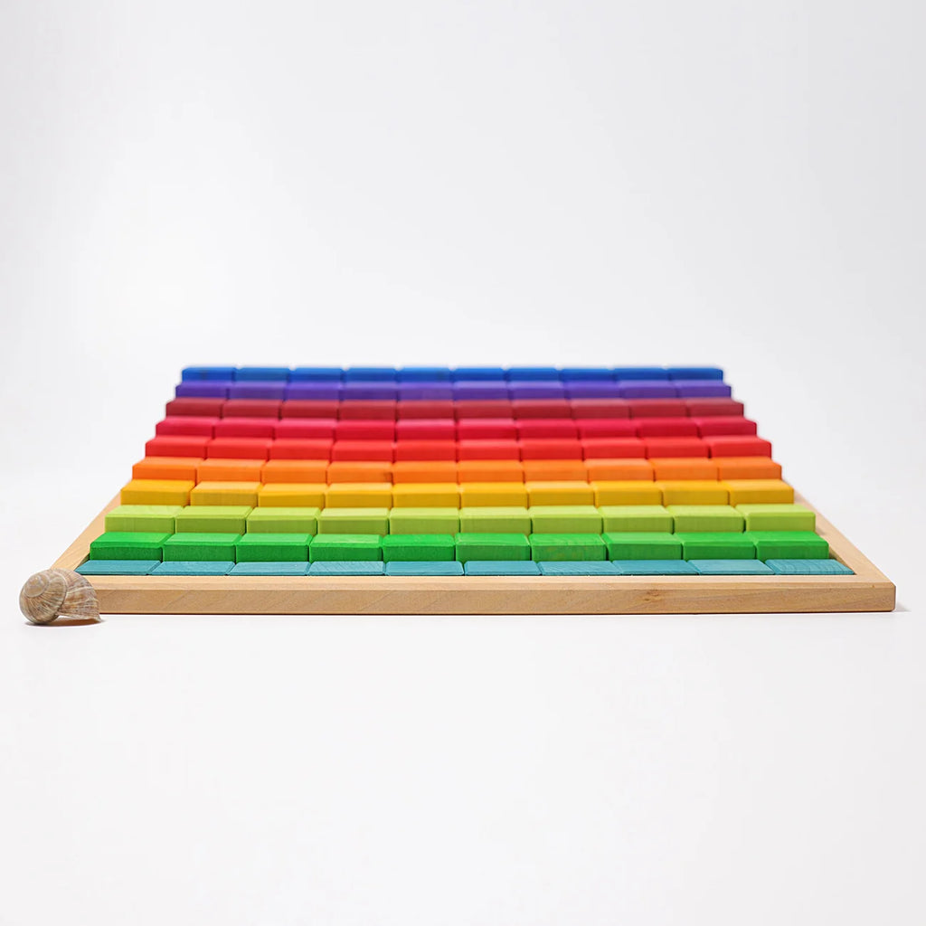 A wooden tray of Grimm’s Stepped Counting Blocks - Large, featuring rows of rainbow-colored wooden building blocks arranged by height with a single snail shell in the foreground.