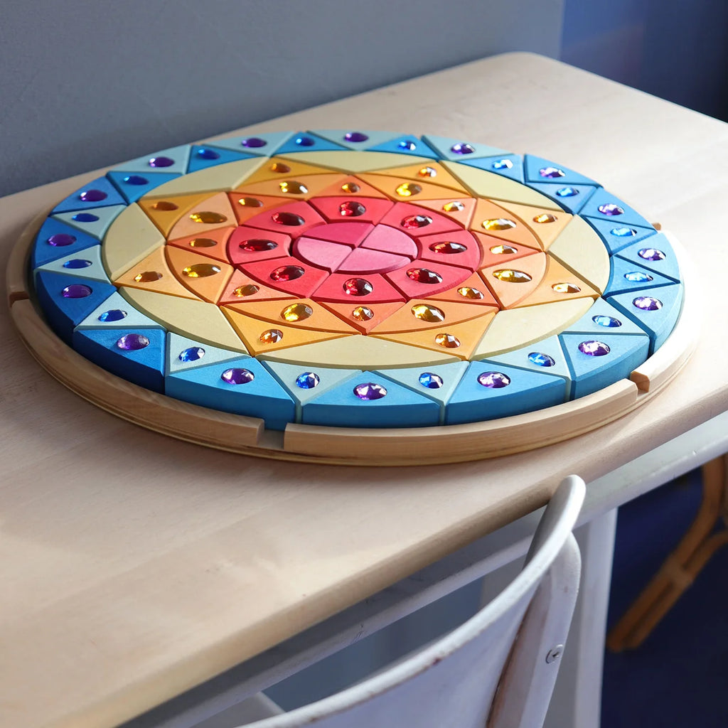 A circular wooden tray holds the Grimm’s Sparkling Mandala Sun Creative Puzzle, featuring geometric triangles and diamonds inlaid with reflective, jewel-toned gems to support fine motor skills.