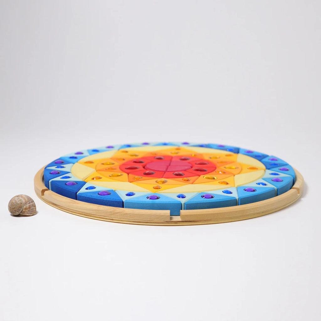 A circular wooden tray holds a colorful Grimm’s Sparkling Mandala Sun Creative Puzzle made of geometric blocks embedded with gemstone-like accents, next to a small snail shell.