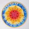 The Grimm’s Sparkling Mandala Sun Creative Puzzle features a circular wooden frame filled with geometric blocks in a radial gradient of red, orange, and yellow, accented with blue triangular pieces and embedded faceted jewels.