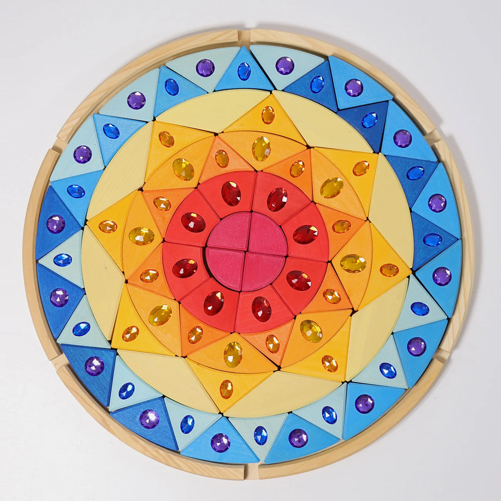 The Grimm’s Sparkling Mandala Sun Creative Puzzle features a circular wooden frame filled with geometric blocks in a radial gradient of red, orange, and yellow, accented with blue triangular pieces and embedded faceted jewels.