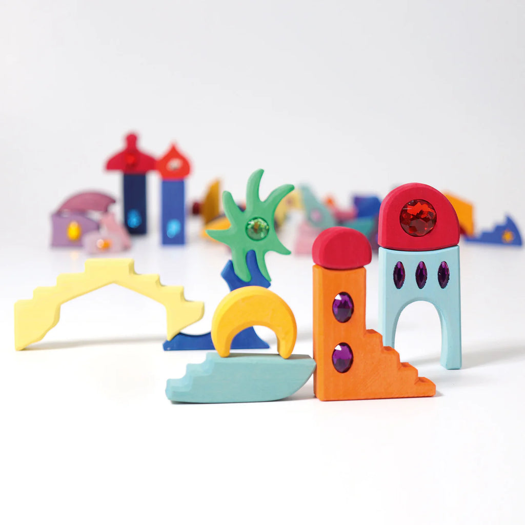 Grimm's Sparkling Mandala Orient wooden blocks, featuring arches, steps, and towers with jewel-like glitter accents, are artfully arranged to create an abstract city on a white background.