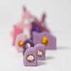 A row of small pastel purple and pink glittery wooden blocks with gem-like decorations from Grimm's Sparkling Mandala Orient (ships in 1-2 weeks) is arranged against a white background, evoking imaginative play.