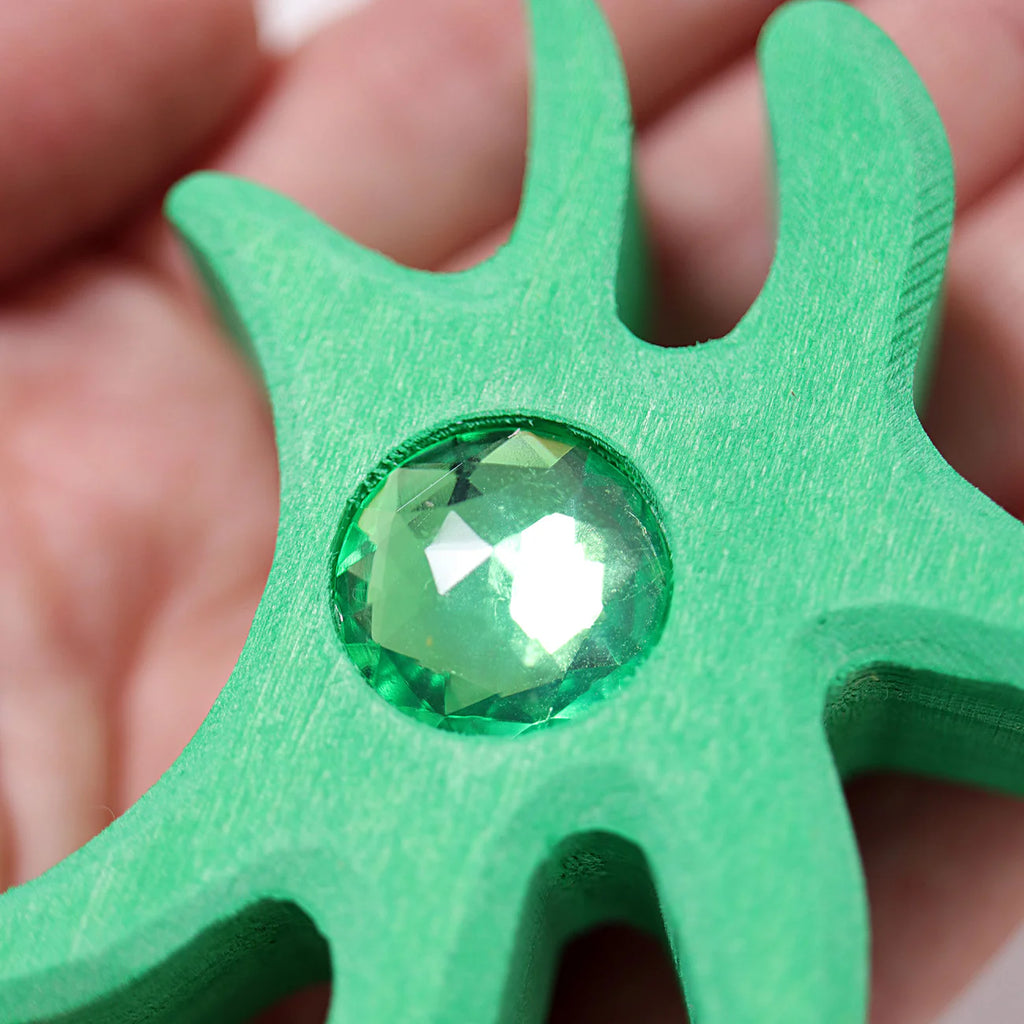 A hand holds Grimm's Sparkling Mandala Orient, a green abstract-shaped piece with curved extensions, featuring a large faceted green gemstone and glitter stones at its center (ships in 1-2 weeks).