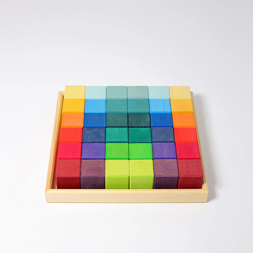 The Grimm's Rainbow Mosaic Building Blocks are arranged in a wooden tray, featuring vibrant blocks in a beautiful gradient of yellow, blue, green, red, and purple to create a striking rainbow pattern. Perfect for developing fine motor skills and spatial thinking, the set is displayed on a white background.