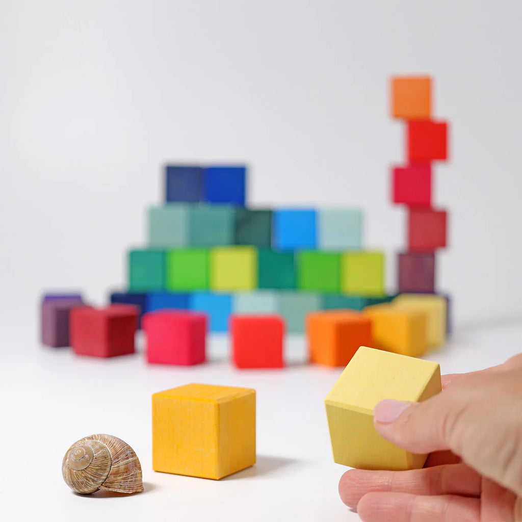 A hand holds a yellow block from Grimm's Rainbow Mosaic Building Blocks near a snail shell, engaging fine motor skills, while the colorful Grimm's blocks are arranged in the background to encourage spatial thinking.