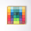 A wooden tray displays the Grimm's Rainbow Mosaic Building Blocks, featuring vibrant blocks arranged in a gradient pattern. The colors transition from yellow, orange, and blue at the top to green, purple, and red at the bottom, all set against a white background. This engaging set is ideal for fostering fine motor skills and spatial thinking in children.