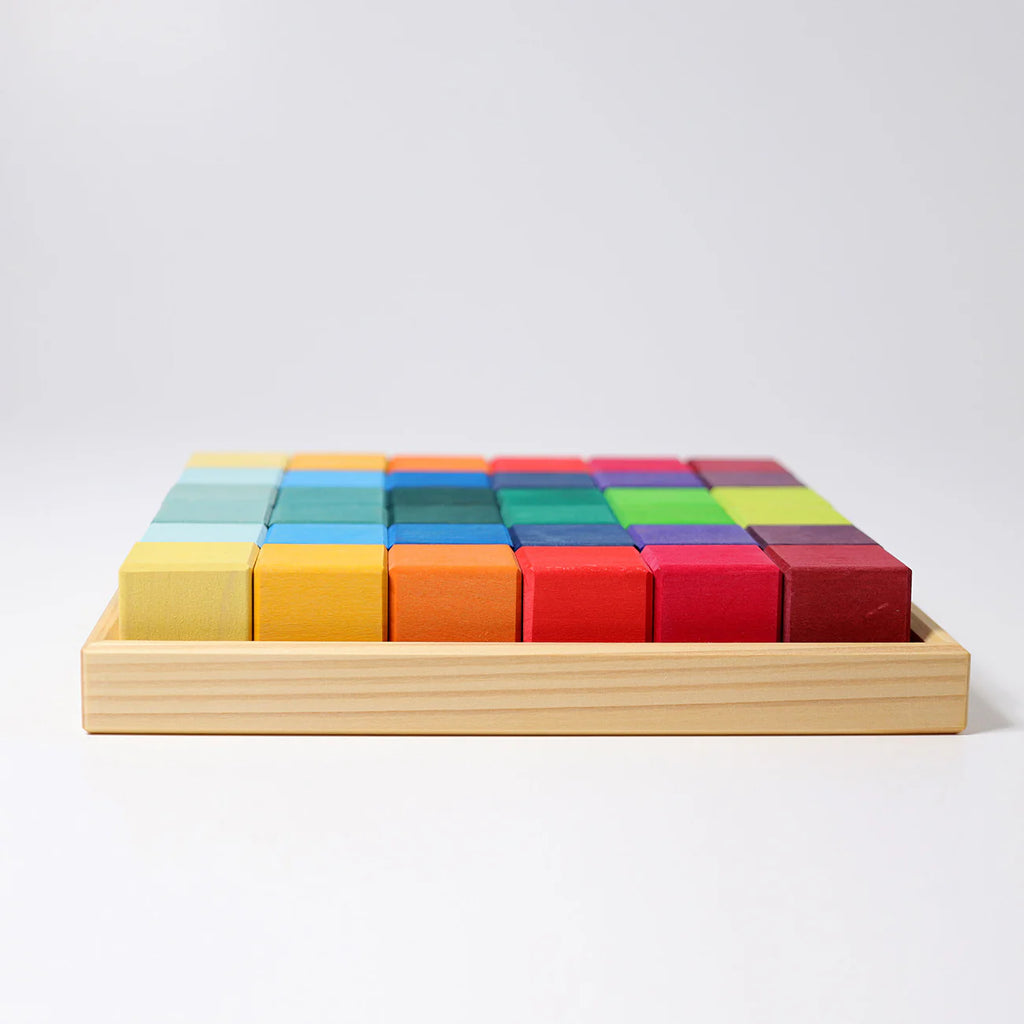 A wooden tray holds neatly arranged Grimm's Rainbow Mosaic Building Blocks in rows, featuring shades of yellow, orange, blue, green, red, and purple against a plain white background—perfect for encouraging spatial thinking skills and fine motor development.