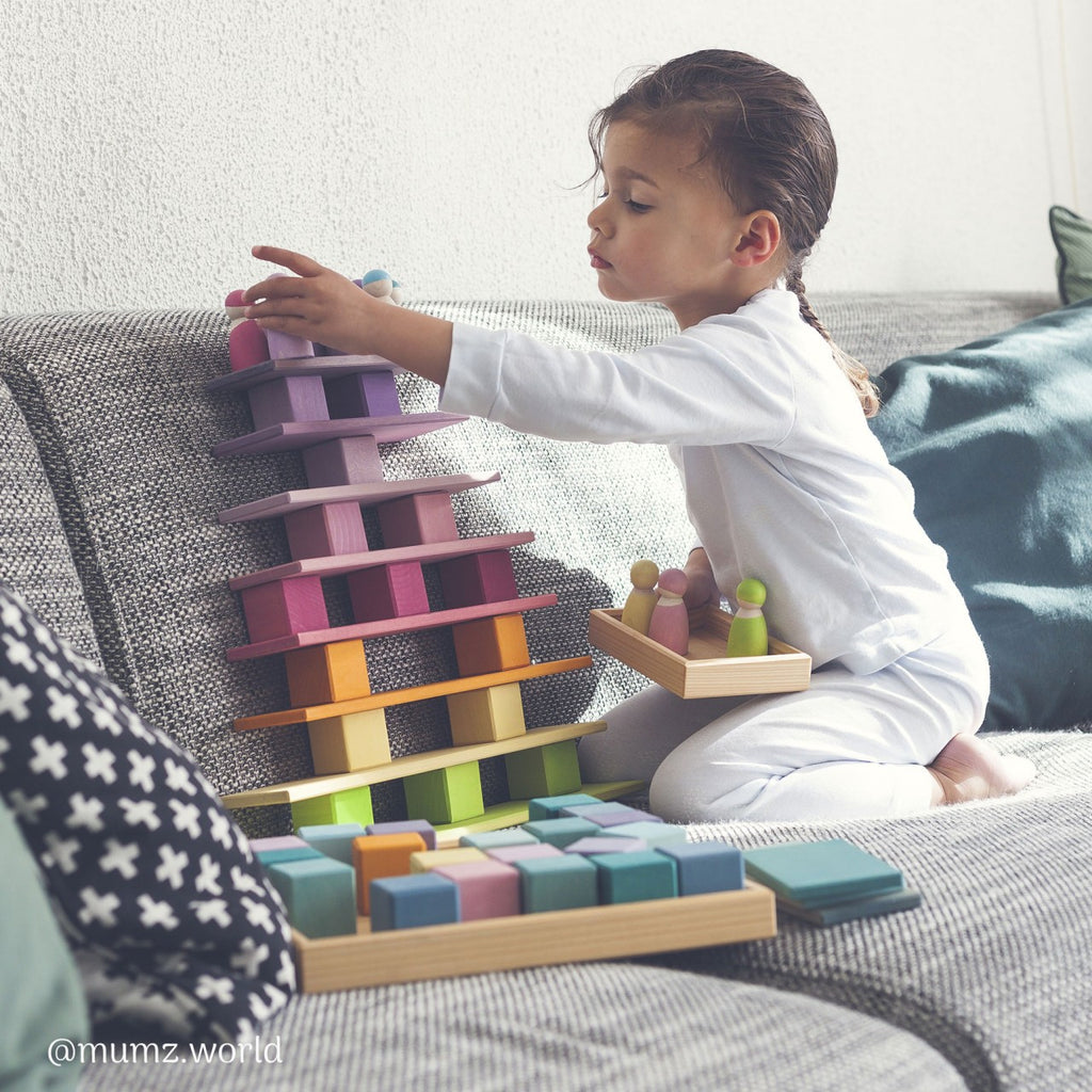 A young child in white pajamas sits on a gray couch, focused on playing with building blocks. The Grimm's Pastel Mosaic Building Blocks, made of pastel-colored blocks arranged in a triangular tower shape, helps develop fine motor skills. Cushions adorn the couch, and the background is light-colored.