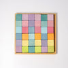 A wooden tray holding 36 Grimm's Pastel Mosaic Building Blocks arranged in a 6x6 grid. The blocks come in various pastel shades of purple, blue, green, pink, yellow, and orange, creating a vibrant and symmetrical pattern. Ideal for children to enhance fine motor skills against the plain white background.