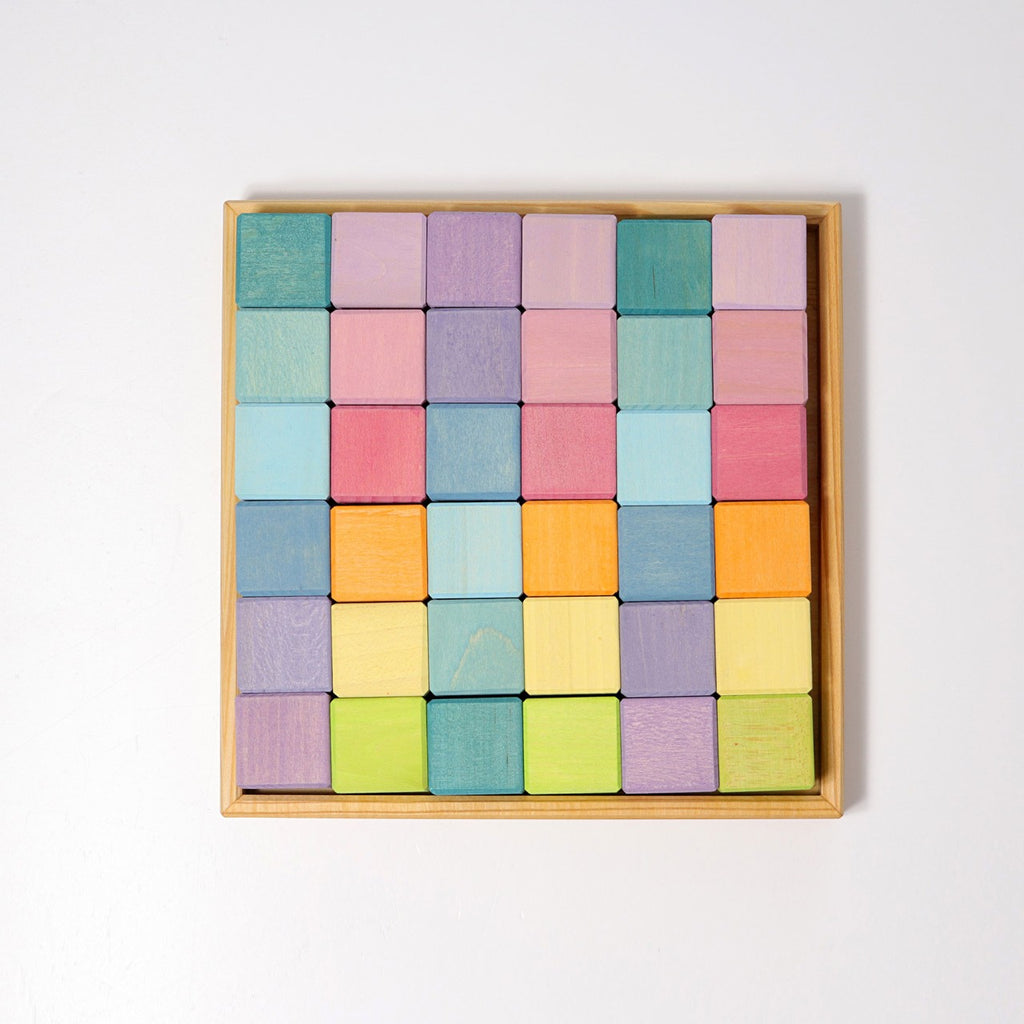 A wooden tray holding 36 Grimm's Pastel Mosaic Building Blocks arranged in a 6x6 grid. The blocks come in various pastel shades of purple, blue, green, pink, yellow, and orange, creating a vibrant and symmetrical pattern. Ideal for children to enhance fine motor skills against the plain white background.