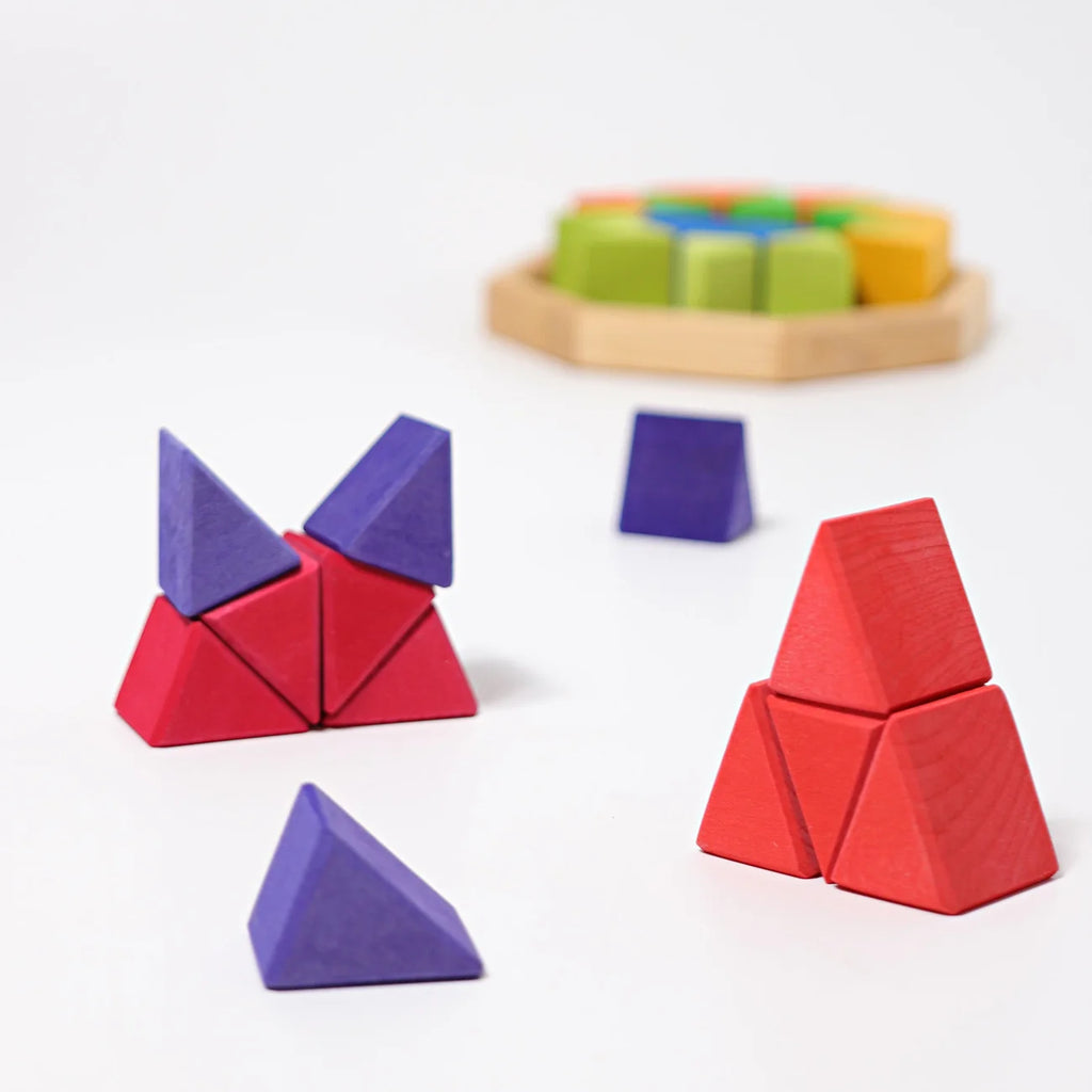 Grimm’s Medium Octagon (ships in 2-3 weeks) features colorful wooden geometric building blocks, such as red triangles and purple pyramids, arranged in small structures on a white surface, with a wooden frame tray of additional blocks visible blurred in the background.