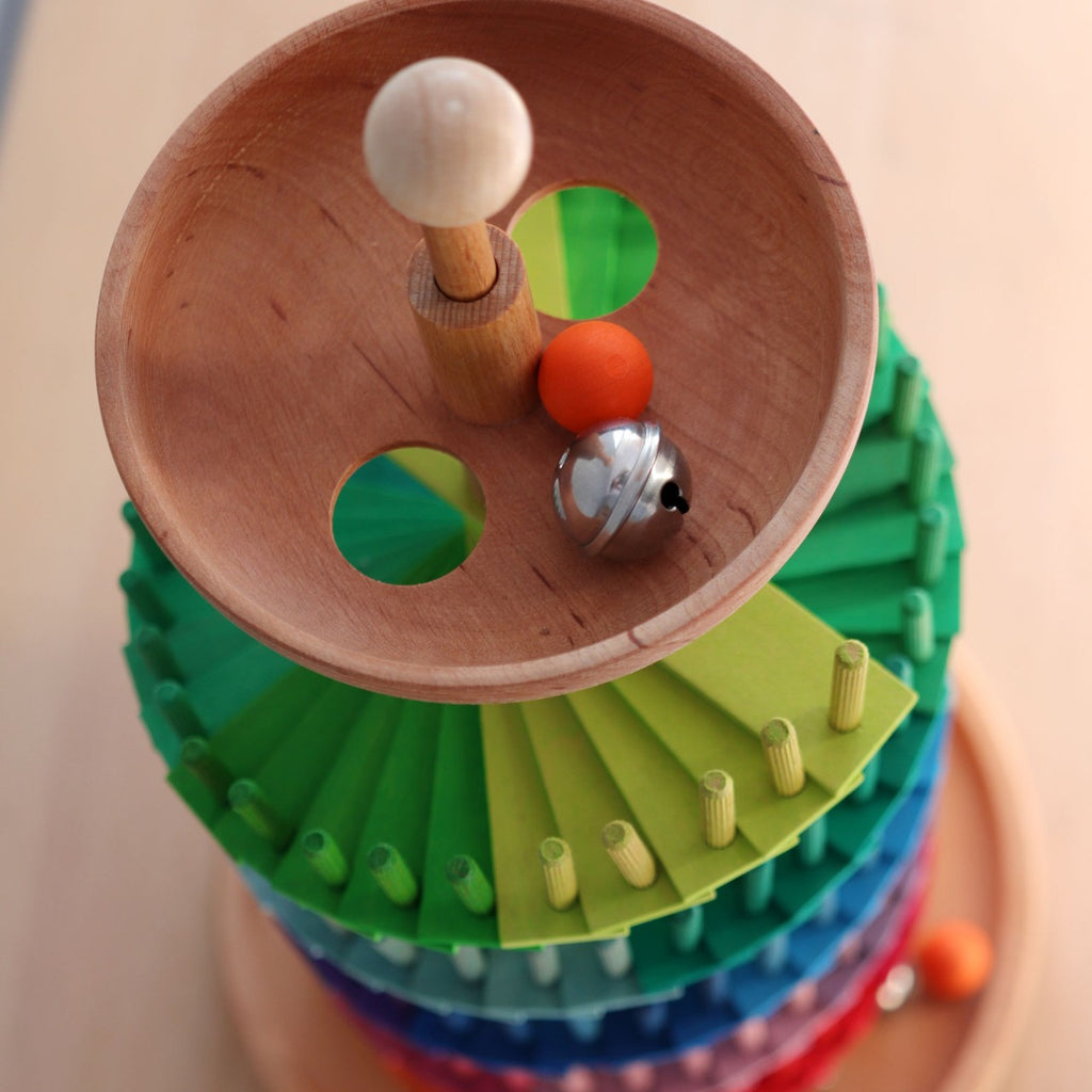 The Grimm's Assembly Set Musical Ball Run features colorful wooden ramps, a wooden ball, an orange ball, and a silver bell on top. Viewed from above, two holes in the top bowl make this an engaging educational toy for kids.