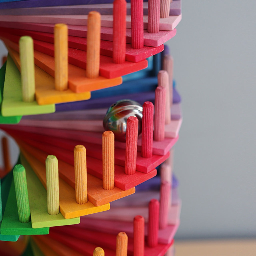 Close-up of Grimm's Assembly Set Musical Ball Run: stacked, spiraling rainbow platforms with a silver marble rolling down. This colorful educational toy features vertical pegs and a blurred background.