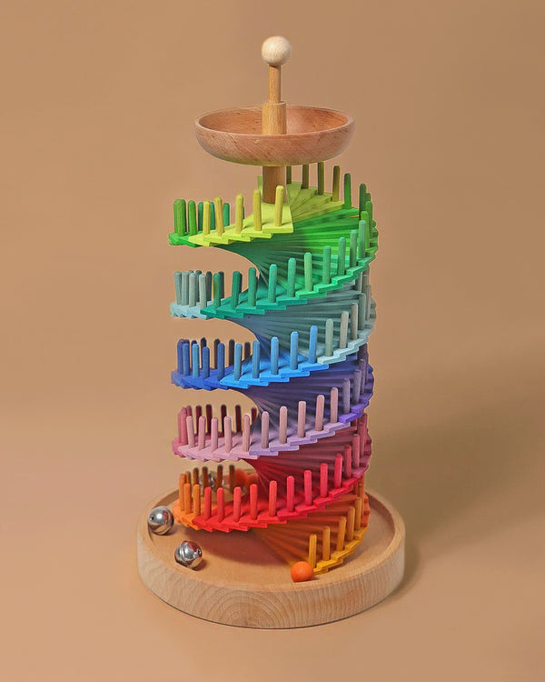 Grimm's Assembly Set Musical Ball Run features a rainbow spiral track with vertical pegs. Two metallic marbles and one orange marble sit on a wooden tray, making it a colorful and engaging construction toy for kids.