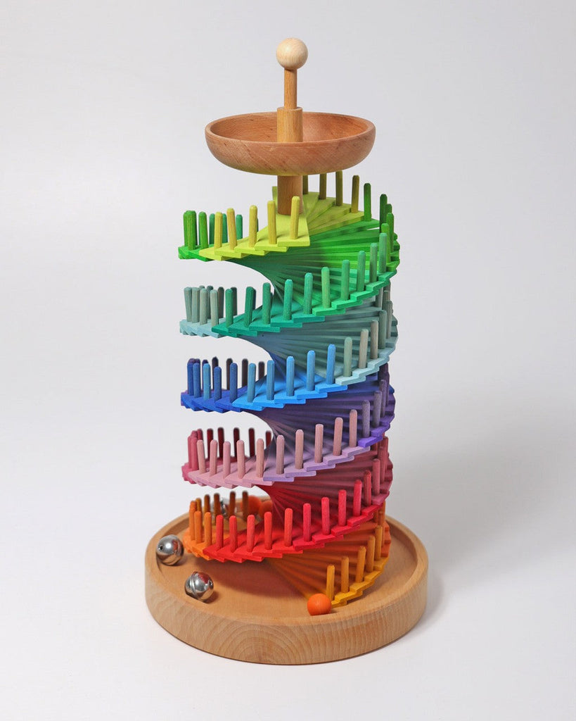 The Grimm's Assembly Set Musical Ball Run features wooden, spiraling colorful tracks and rainbow pegs topped with a wooden bowl. Three marbles—one orange and two metallic—rest at the base of this engaging educational toy.