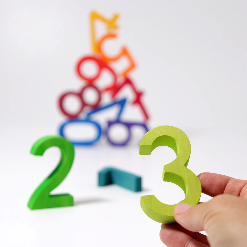 A hand holds a green number three from Grimm's Building Set - Numbers, with a green two and blue one nearby. Blurred in the background are stacked colorful wooden numbers, perfect for building, stacking, and motor skill development.