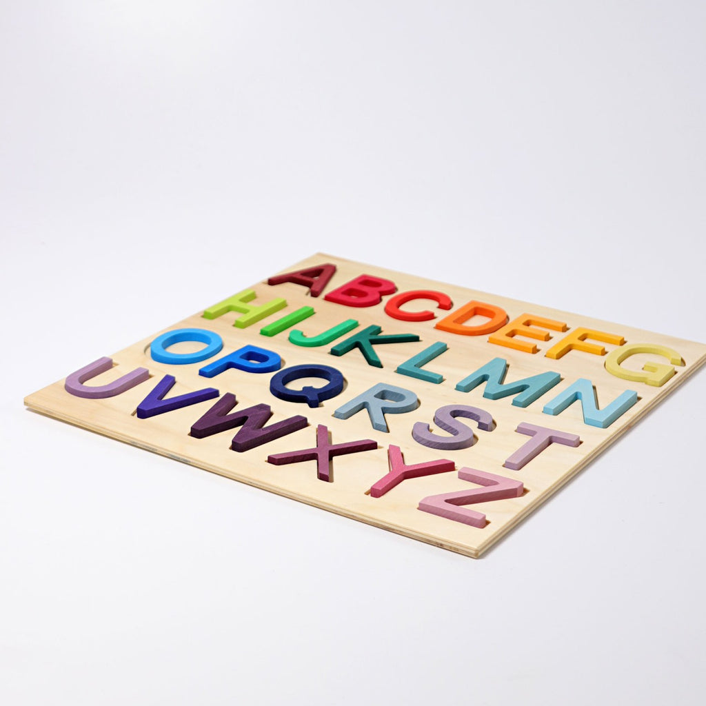 The Grimm's ABC Game is a wooden alphabet puzzle showcasing vibrant, three-dimensional letters arranged sequentially. Each letter is painted in a unique color from the palette of red, blue, green, yellow, and purple. The puzzle board features a light wood hue with cutout slots for each letter and is crafted within a robust wooden frame made from non-toxic materials.