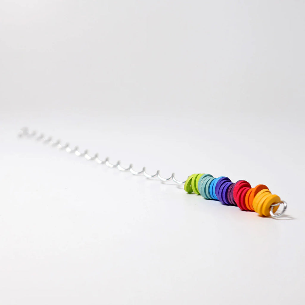 The Grimm's Spirelli Clatter Wand, a spiral metal rod adorned with vibrant wooden beads stacked in rainbow sequence at one end—evoking the look of cascading rings from a classic wooden spiral toy—rests against a plain white background.