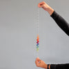 Two hands hold the Grimm's Spirelli Clatter Wand vertically. In the center, cascading rings create a colorful, spiral-shaped design in a rainbow gradient against a plain gray background.