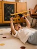 A child lies on the Little Forest Animals — The Aviator polka dot rug, holding a handcrafted metal biplane overhead, with a chalkboard drawing and wooden furniture in the background.