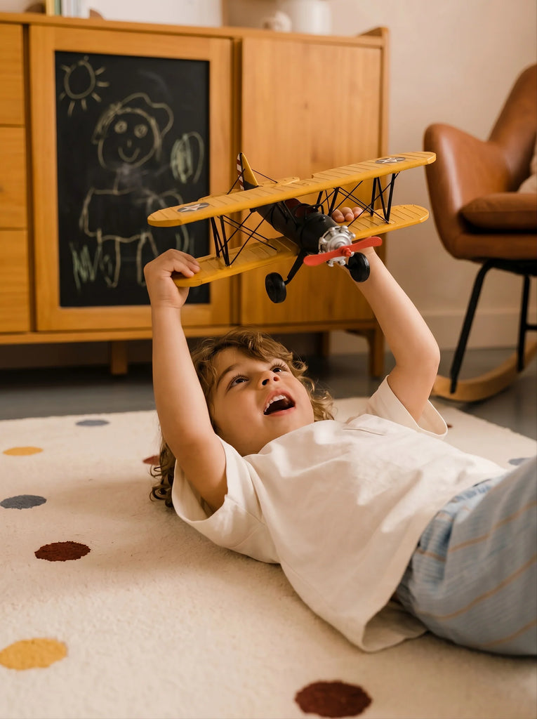 A child lies on the Little Forest Animals — The Aviator polka dot rug, holding a handcrafted metal biplane overhead, with a chalkboard drawing and wooden furniture in the background.
