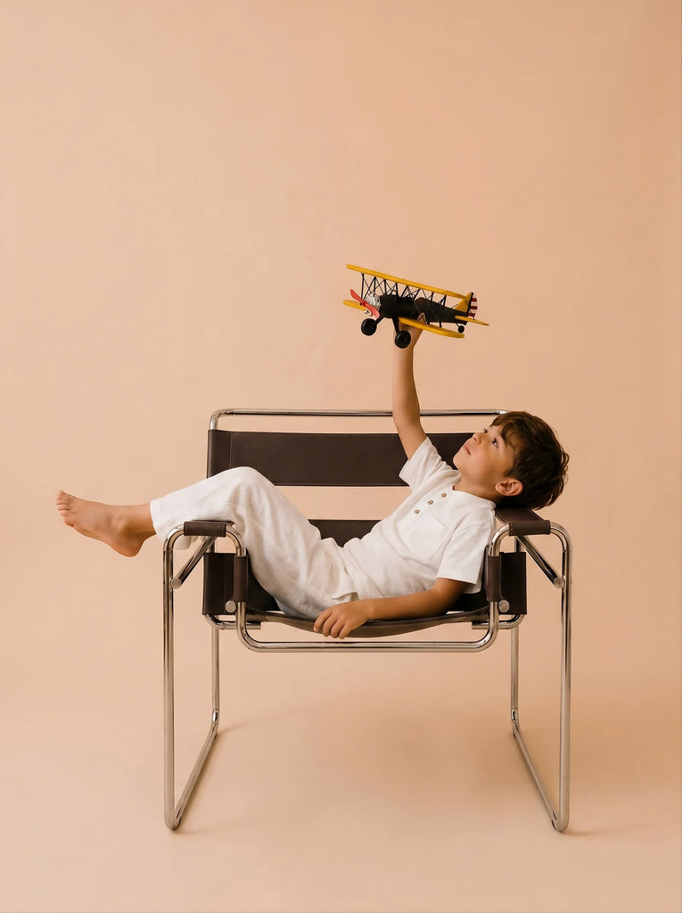 A young boy in white clothes reclines on a modern chair, holding Little Forest Animals — The Aviator handcrafted metal biplane above him against a beige background.