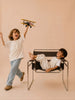 A smiling child walks while holding Little Forest Animals — The Aviator, a handcrafted metal biplane. Another child lounges in a modern chair, watching. Both are set against a neutral beige background.