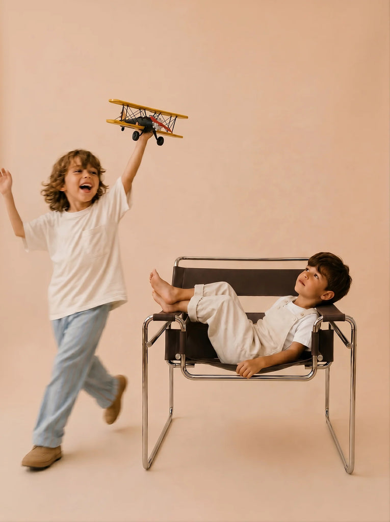 A smiling child walks while holding Little Forest Animals — The Aviator, a handcrafted metal biplane. Another child lounges in a modern chair, watching. Both are set against a neutral beige background.