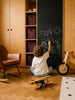 A child sits on the floor drawing with chalk on a blackboard wall—sketching a plane, sun, stick figure, and house—in a cozy playroom filled with wooden furniture and shelves. Above, Little Forest Animals — The Aviator handcrafted metal biplane adds whimsy as barnstormer plane decor, inspiring endless adventures and creativity.