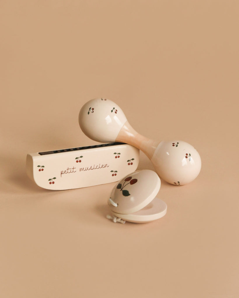 The Konges Sloejd Petit Music Set - Glossy Cherry features three cream-colored wooden instruments with cherry designs—a maraca, a harmonica case labeled "petit musicien," and a clamshell castanet—ideal for any kids’ music set.
