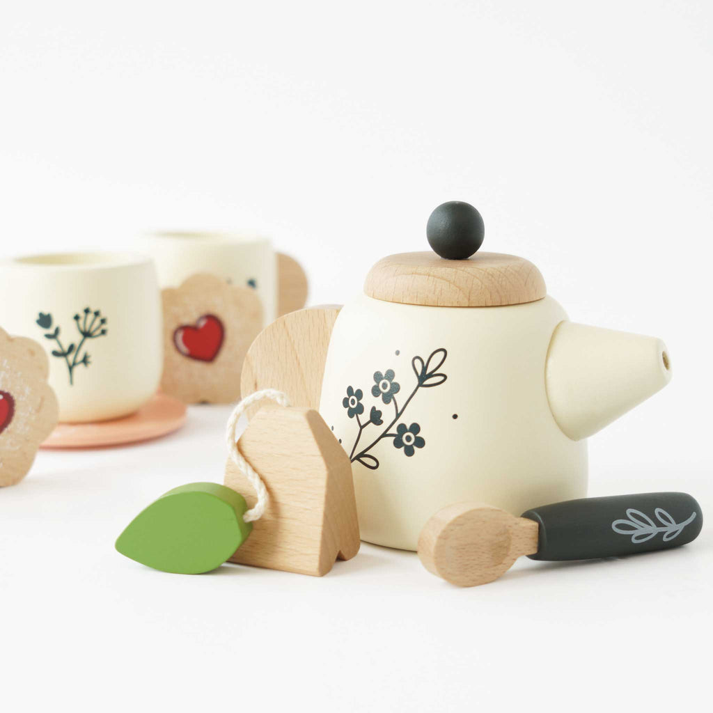 Introducing the Wooden Tea Set & Tray, an ideal choice for imaginative playtime. This eco-friendly set features a charming teapot with a floral design, two cups decorated with heart motifs, and flower-shaped coasters. It also includes a tea bag with a green leaf tag and a small spoon, all beautifully presented on a light-colored surface.