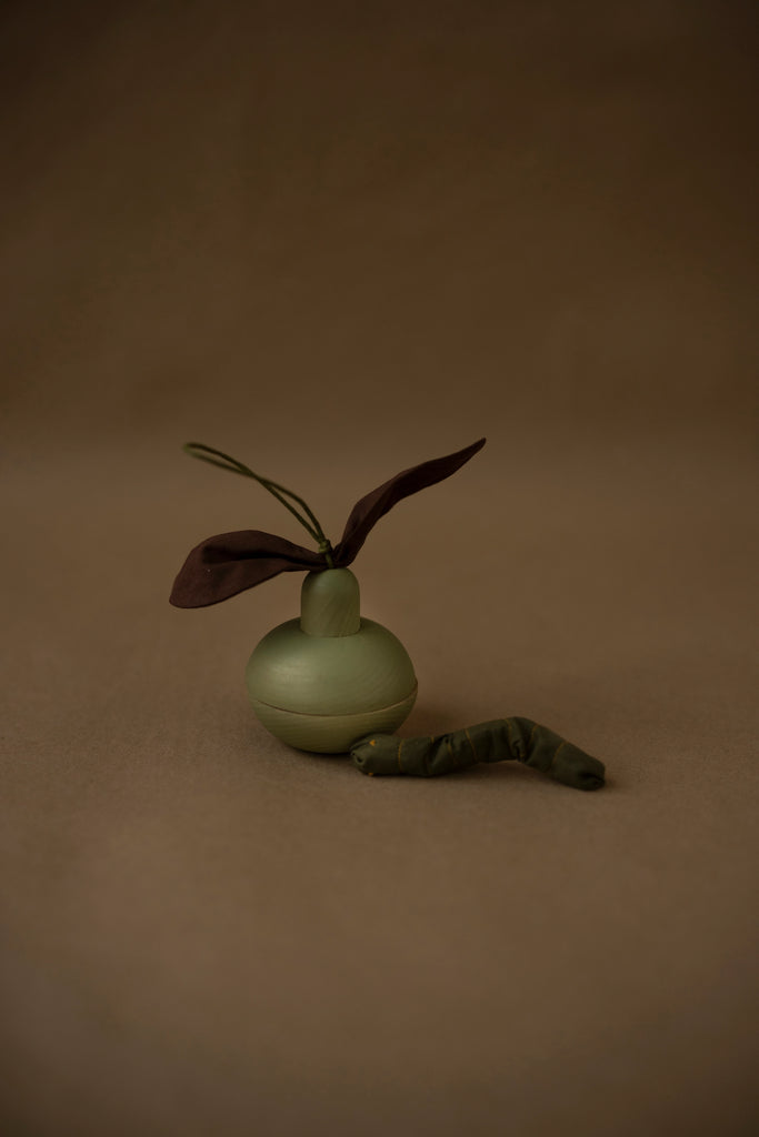 The Wooden Pear & Little Friend set features a small, round wooden pear with a brown leaf-like attachment on top, resting on a brownish background. Accompanying it is a smaller, loosely coiled cotton caterpillar. The overall lighting is muted, giving the scene an earthy tone.