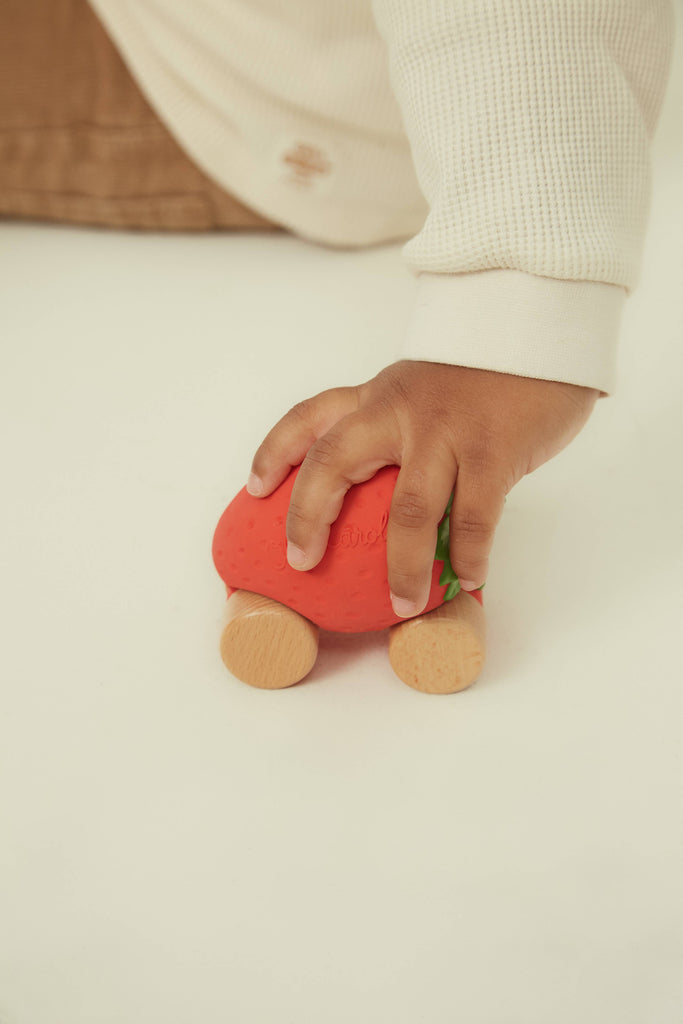 A child in a cream sweater holds Sweetie The Strawberry Baby Car, an eco-friendly red strawberry-shaped toy with wooden wheels, on a white surface.