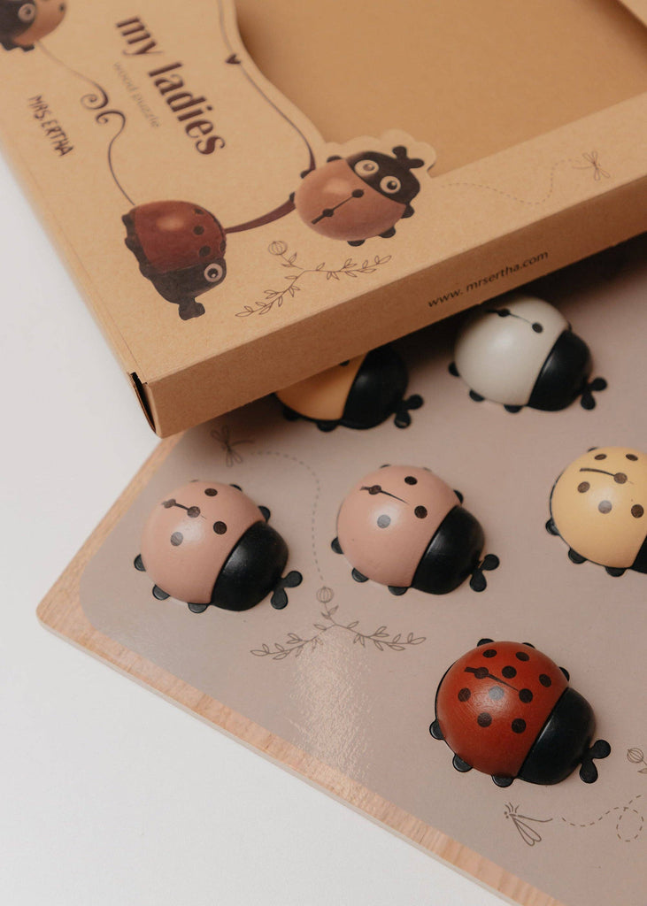 An open box labeled “My Little Lady Bug Wooden Puzzle” reveals a board and six ladybug-shaped game pieces in a variety of colors, such as red, beige, and black. The whimsical design adds a playful element to this counting toy, making it a charming addition to any collection of Montessori toys.