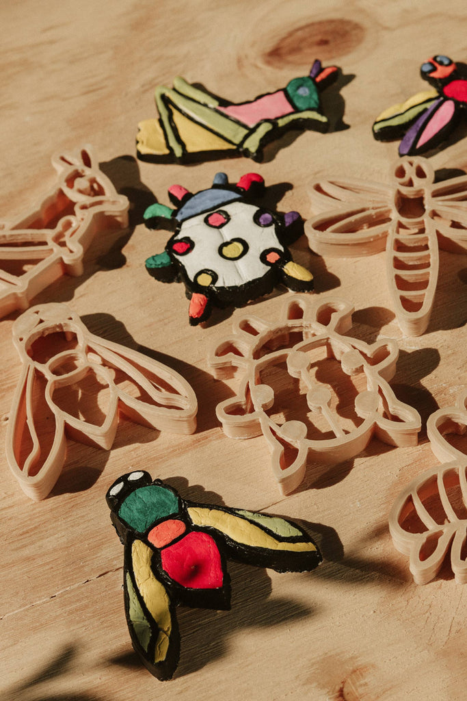 The Insect Cutter Set - Eco Playdough Tool features colorful clay insects and bugs surrounded by matching beige plastic insect playdough cutters, all arranged on a wooden surface in warm sunlight.
