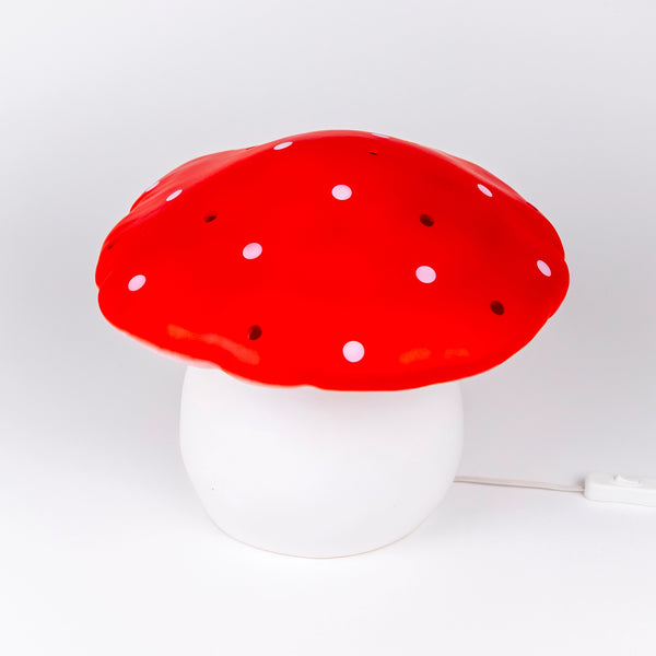 The Red Mushroom Lamp is a whimsical night light with a white stem and red cap topped with white spots, ideal for adding charm to any child's room or nursery. Displayed on a plain white background.