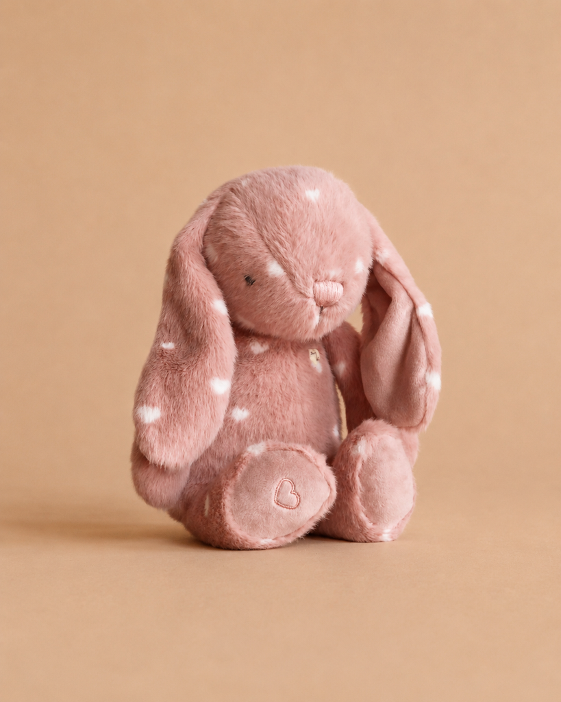 Snuggle Bunnies - Penelope - Little Love, a blush pink bunny soft toy with floppy ears and white spots, sits on a matching tan background. A small heart is stitched on its paw, making it a charming keepsake gift.