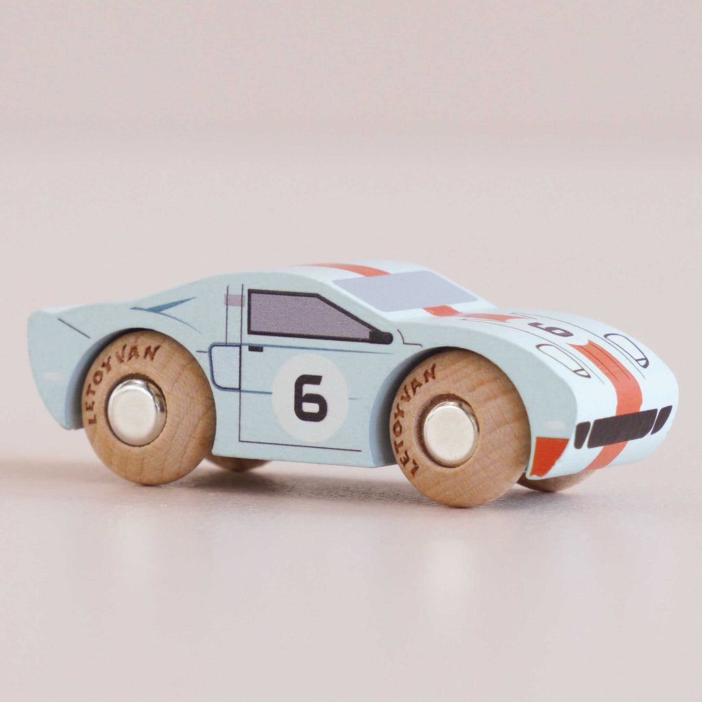 A No. 26 GT145 Fred wooden racing car, crafted in England, featuring a light blue body with the number 6 on the door and hood, black windows, and natural wood wheels, displayed on a light surface.