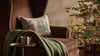 A cozy brown armchair with a green knit blanket and the Coral & Tusk Christmas Tree Sled Pillow sits by a small table with a mug, while a decorated, glowing Christmas tree is visible in the background.