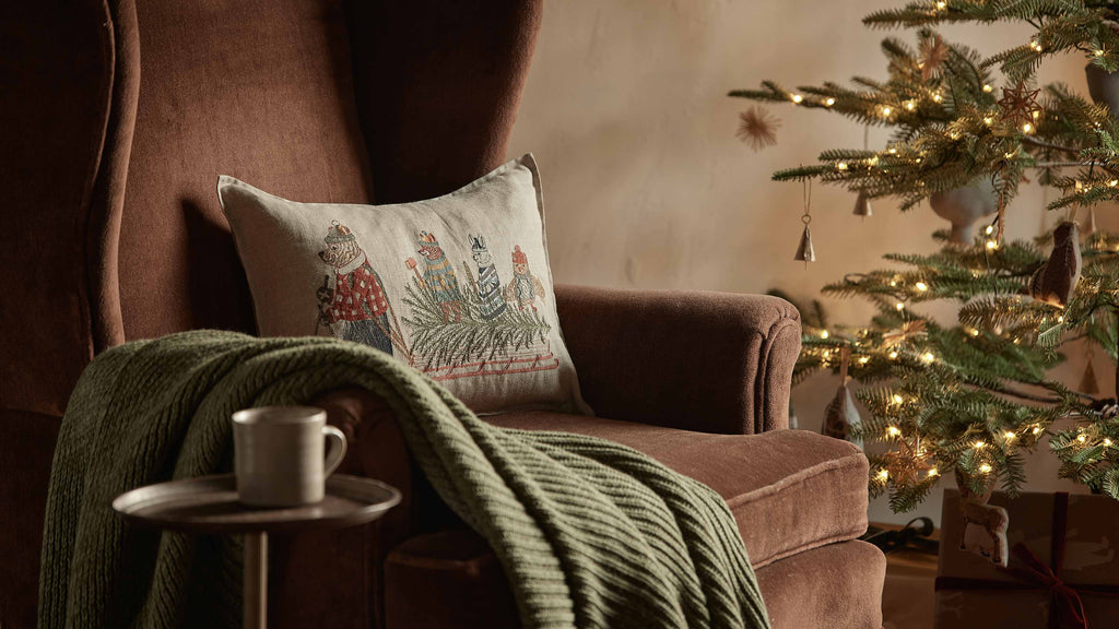 A cozy brown armchair with a green knit blanket and the Coral & Tusk Christmas Tree Sled Pillow sits by a small table with a mug, while a decorated, glowing Christmas tree is visible in the background.