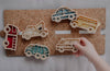 A child’s hand places an ambulance-shaped piece from the Service Vehicle Cutter Set - Eco Playdough Tool onto a corkboard alongside colorful vehicle pieces, such as a bus, police car, fire truck, and tow truck—perfect for inspiring sensory play with this transport-themed playdough cutter set.