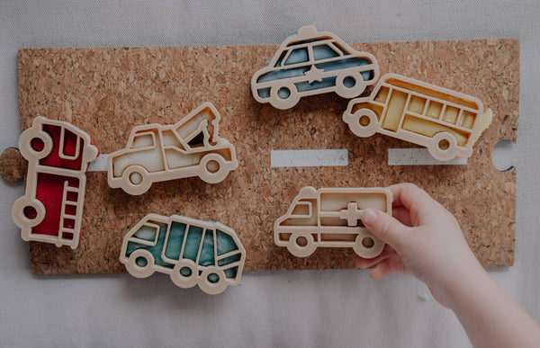 A child’s hand places an ambulance-shaped piece from the Service Vehicle Cutter Set - Eco Playdough Tool onto a corkboard alongside colorful vehicle pieces, such as a bus, police car, fire truck, and tow truck—perfect for inspiring sensory play with this transport-themed playdough cutter set.