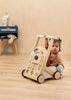 A toddler sits on a wooden floor, holding the Wooden Activity Walker adorned with vibrant shapes and gears. Sensory toys and a stuffed animal rest on a blue table in the background.