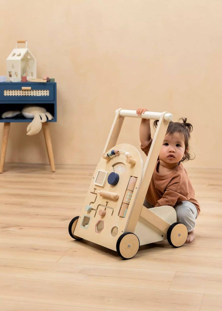 A toddler sits on a wooden floor, holding the Wooden Activity Walker adorned with vibrant shapes and gears. Sensory toys and a stuffed animal rest on a blue table in the background.