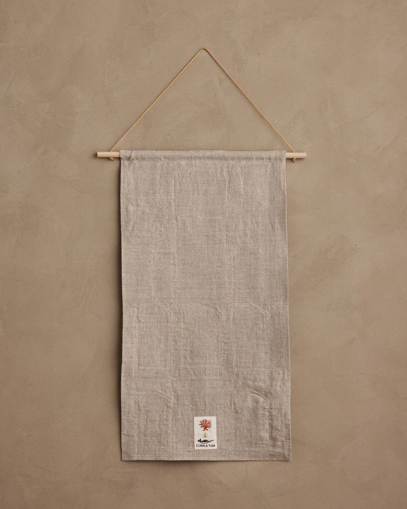 A neutral-colored linen Coral & Tusk Holiday House Advent Calendar suspended from a wooden rod with a thin cord, featuring a small patch at the bottom with an illustration of a coral branch and a fox.