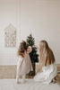 A young girl and a woman with long hair decorate a small Christmas tree in a cozy, minimalist room, placing Sabo Concept Christmas Ornaments amid the white walls and wooden floors.