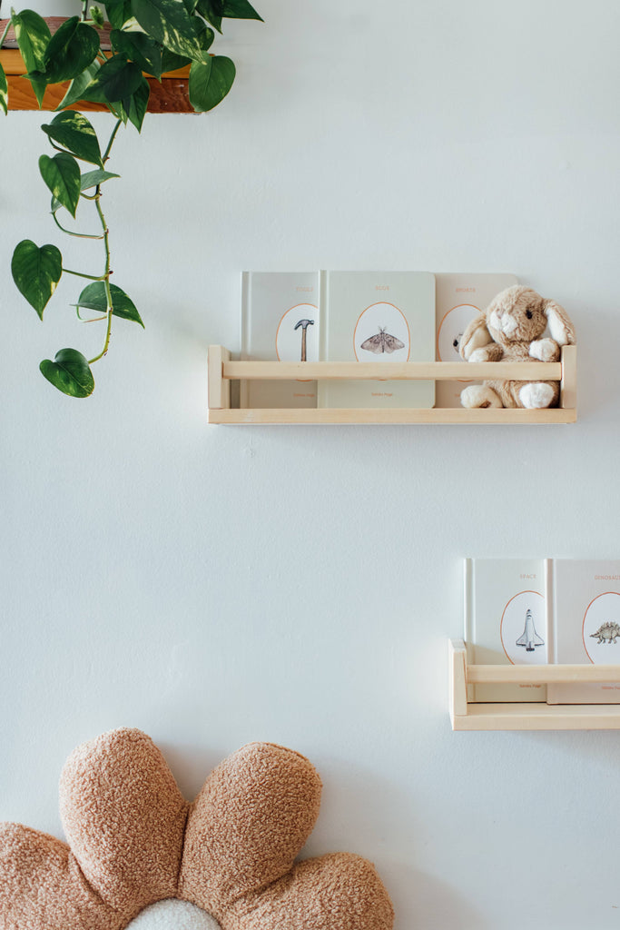 Two wooden shelves with Our Little Library Vol. 3 (5 Board Books) and a plush bunny are on a white wall. A hanging leafy plant and a brown-and-beige flower pillow complete the scene—perfect for early language learning or baby shower gifting.