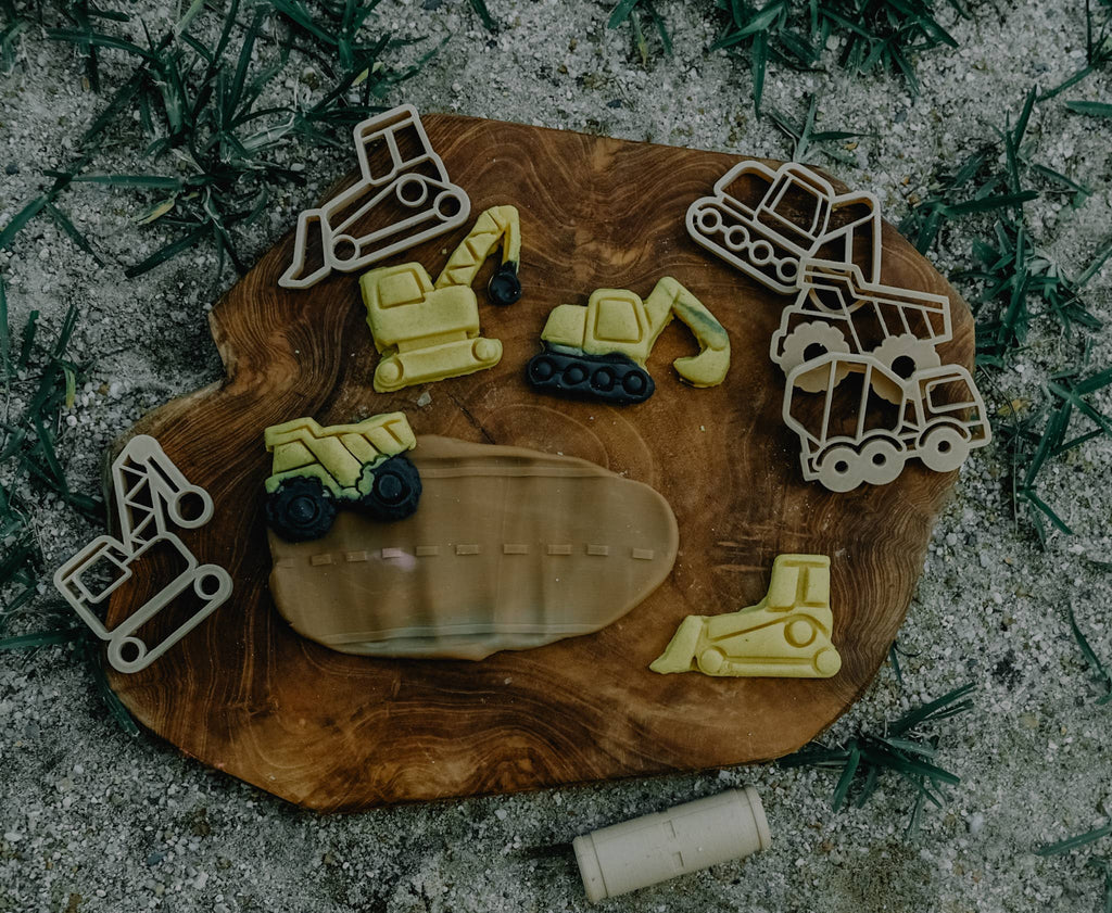 Construction vehicle-shaped cookies and matching Construction Cutter Set - Eco Playdough Tool cookie cutters are displayed on a wooden board atop grass. Accompanied by a small rolling pin and an oval "worksite" slab, this playful scene inspires creative sensory fun—making the Construction Cutter Set - Eco Playdough Tool ideal for imaginative little builders.